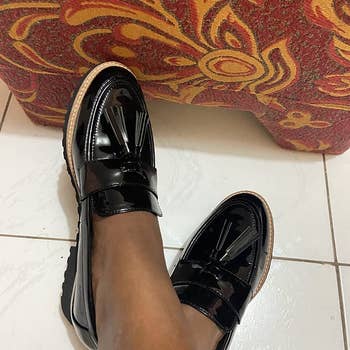 A reviewer photo of a pair of feet wearing the black patent loafers with tassels on the toe 