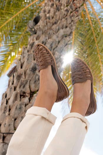 a pair of feet wearing the shoes in dark brown with a palm tree in the background 