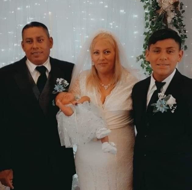 A bride in a white wedding dress and holding an infant stands between a boy and a man in suits and boutonnieres