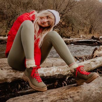 a model wearing a puffer jacket, green leggings, and the boot in khaki green with red laces while sitting on a log 