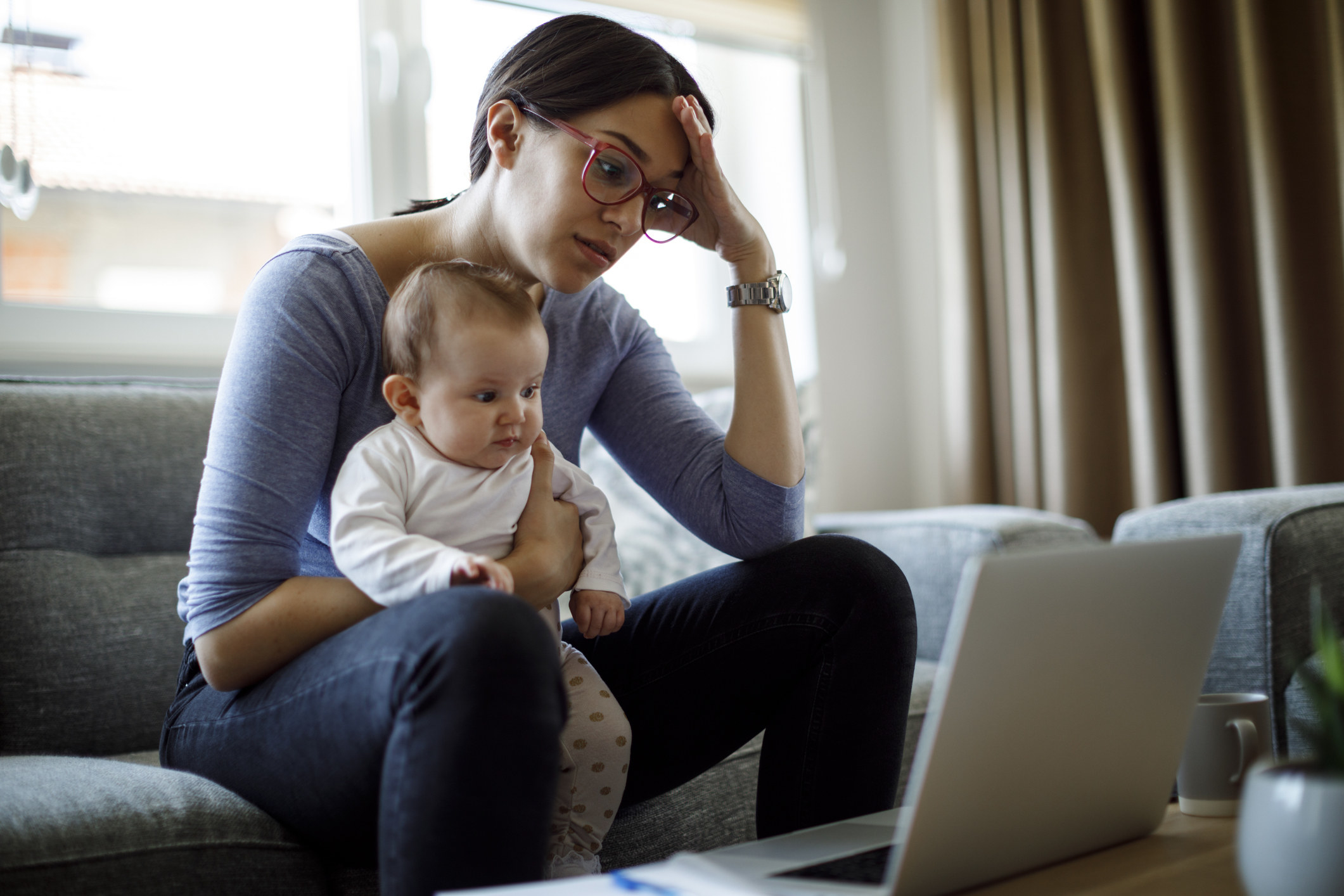 A woman looks tired as she holds a baby and works on her computer