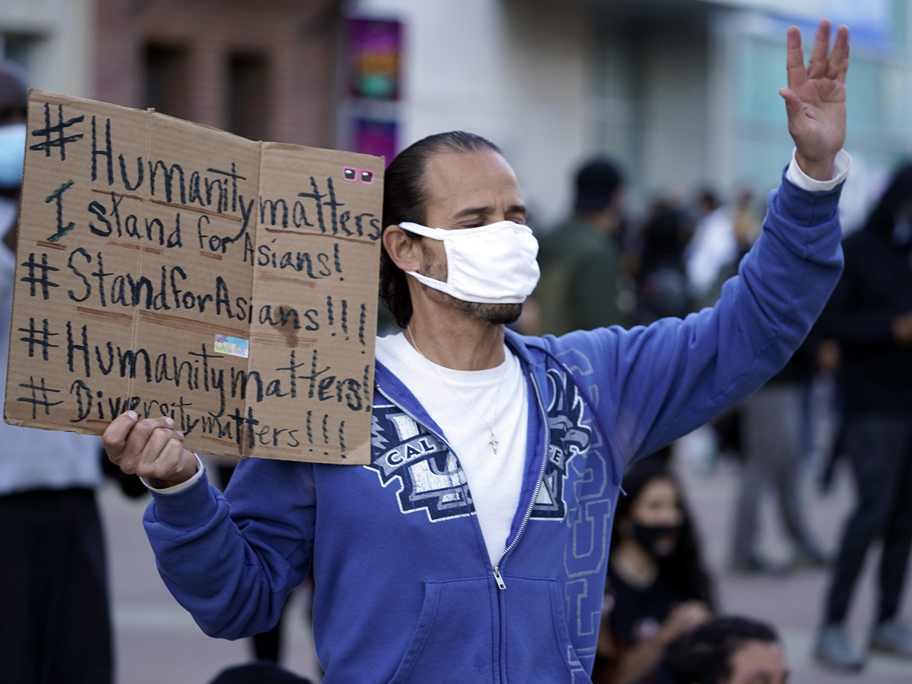 A man holds a cardboard sign that features a series of hashtags, including &quot;humanity matters,&quot; &quot;stand for Asians&quot; and &quot;diversity matters&quot;
