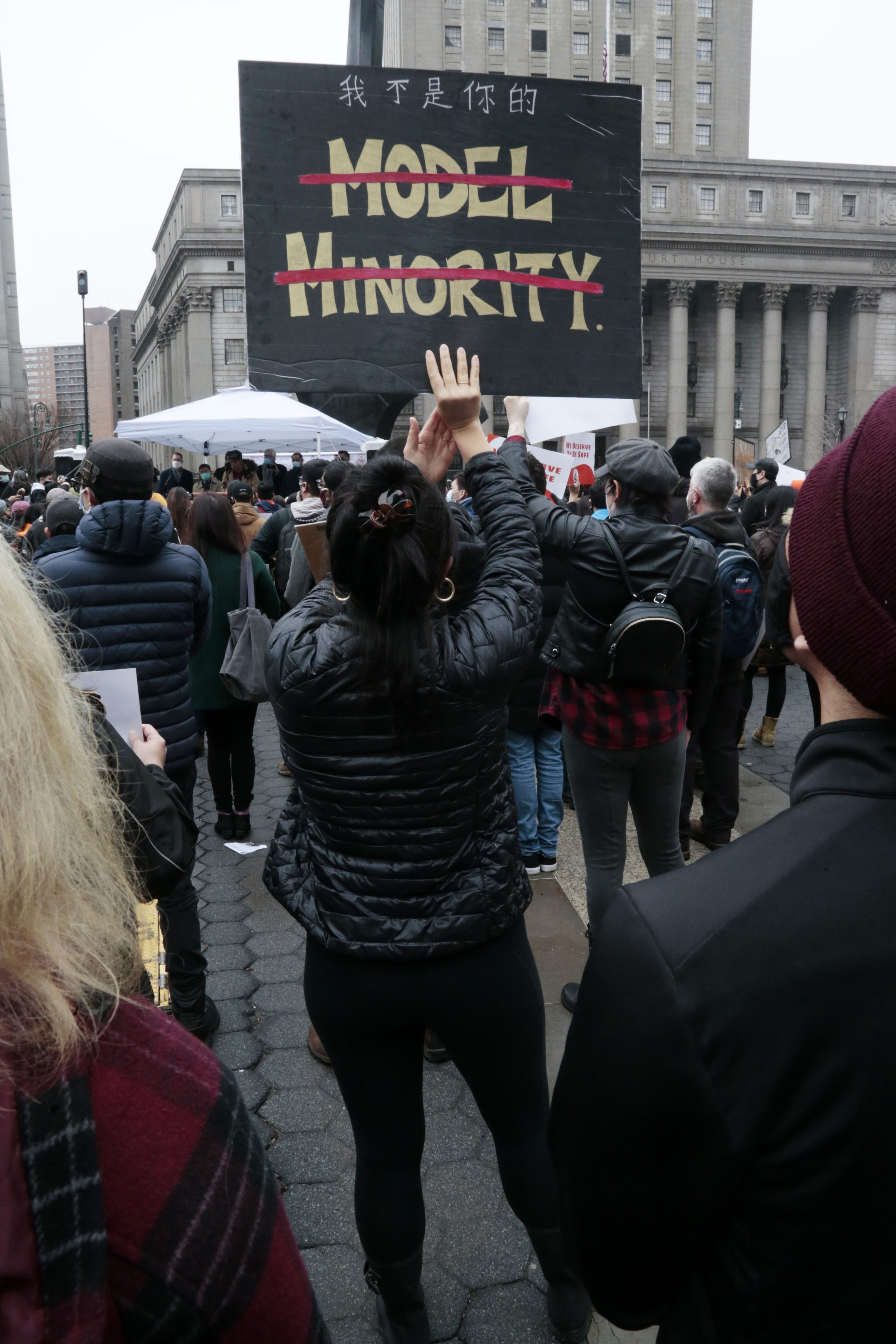 A woman standing with her back to the camera holds a sign with the words &quot;Model Minority&quot; crossed out
