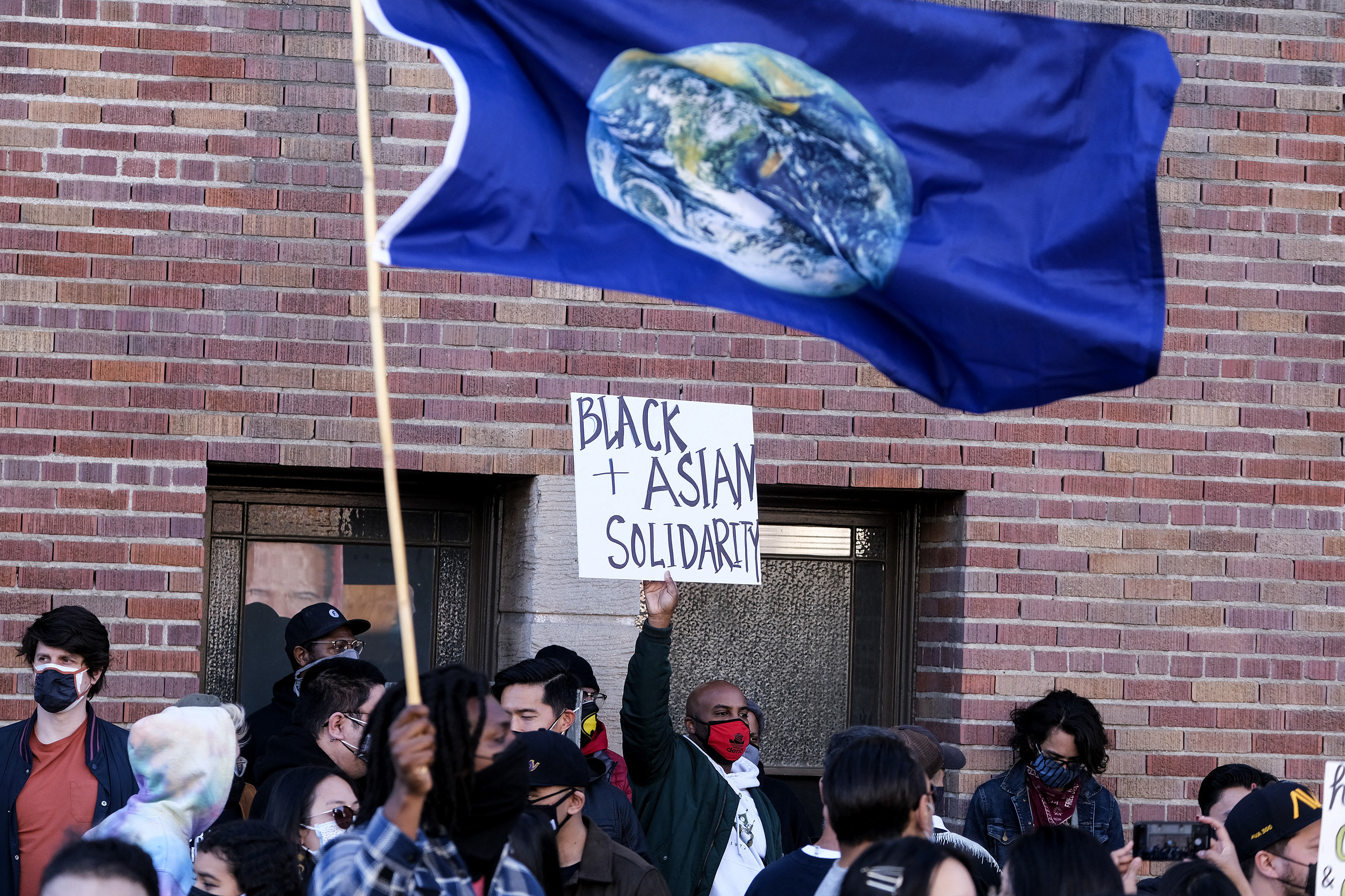 A demonstrator holding a sign for &quot;black &amp; asian solidarity&quot; in a crowd near a person waving a flag with a picture of the Earth