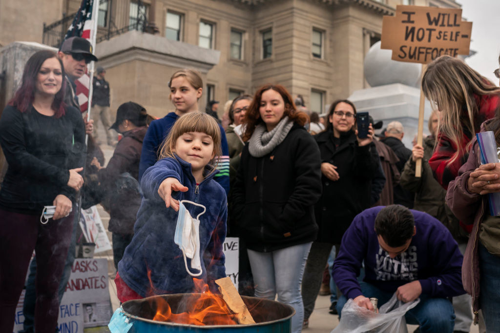A crowd scene showing a young girl throwing a face mask into a garbage can that is on fire