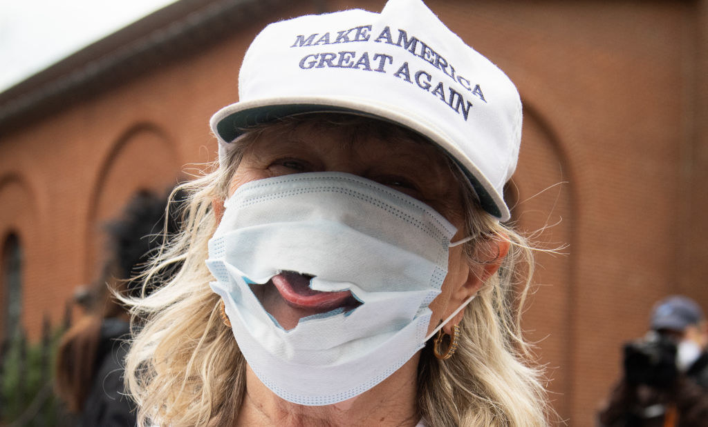 Person wearing a "Make America great again" hat and a face mask with a large hole by the mouth, showing their stuck-out tongue