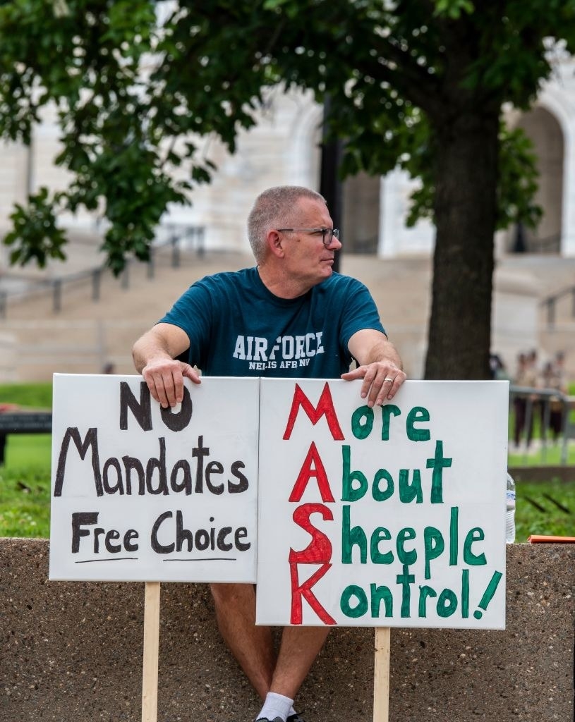 Man in an Air Force T-shirt holding two signs, one reading, "No Mandates Free Choice" and another reading, "More About Sheeple Kontrol!" with the letters M-A-S-K highlighted