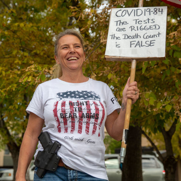 Woman in a "Right to Bear Arms / Girls With Guns" T-shirt and holding a sign reading, "COVID 19-84 the tests are rigged the death count is false"