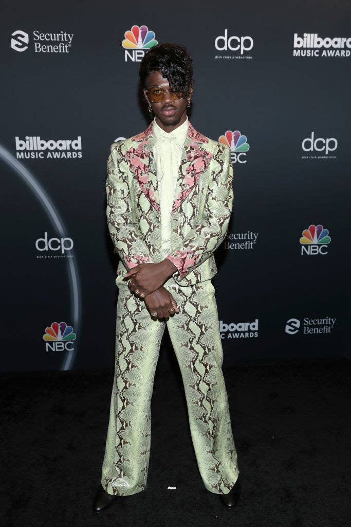 Lil Nas posing on a red carpet in a snakeskin inspired suit, sunglasses, and rocking bangs