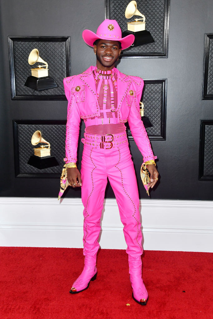 Lil Nas rocking a hot pink suit with cropped jacket, cowboy boots, harness, and hat as well as silk scarves around his wrists