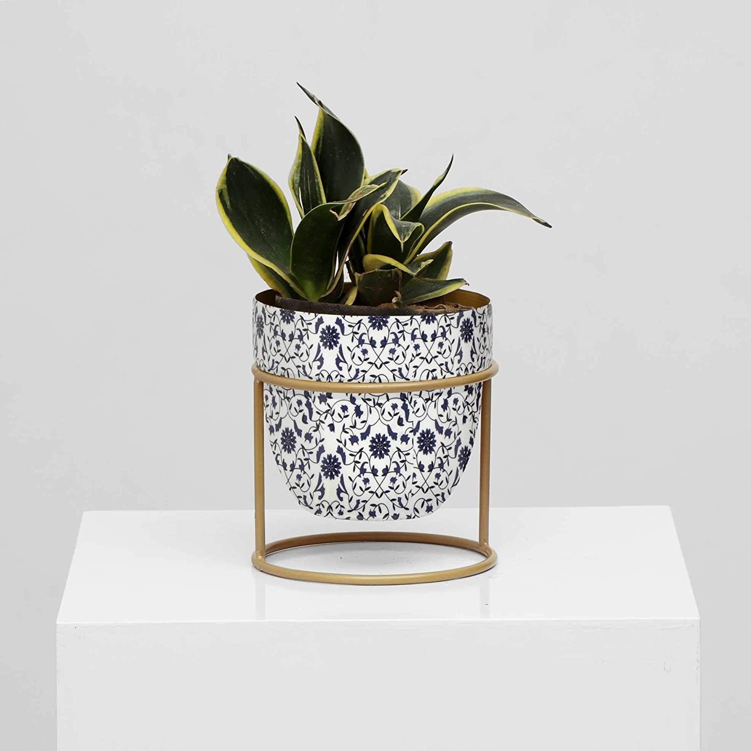 A plant holder in white with a navy blue floral design. It is placed in a golden stand.
