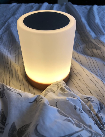 reviewer image of cylindrical small lamp glowing white resting on a bed 