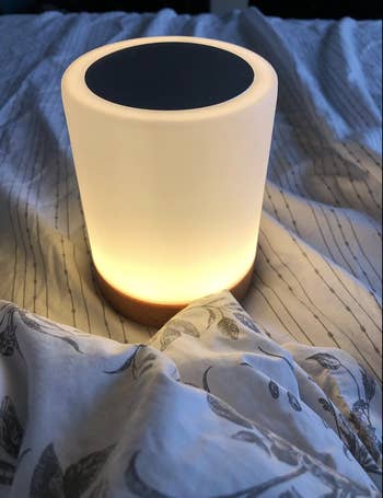 reviewer image of cylindrical small lamp glowing white resting on a bed 