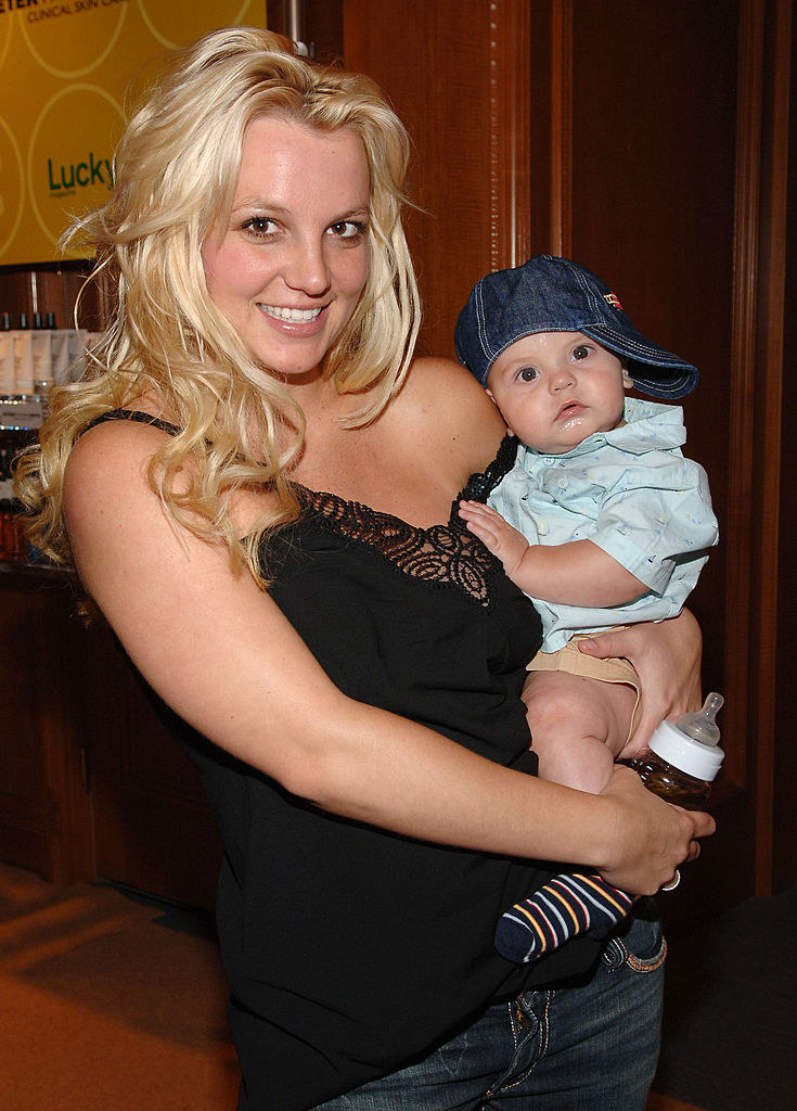 Britney smiling and holding one of her sons as a baby, who looks adorable in his khaki shorts and baseball cap turned to the side