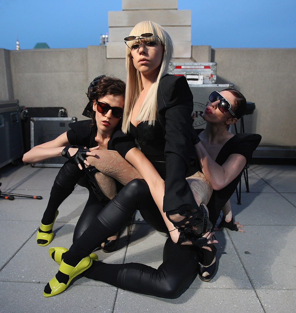 Lady Gaga dramatically posing with two other people for a photo on a rooftop deck as they contort their bodies around each other