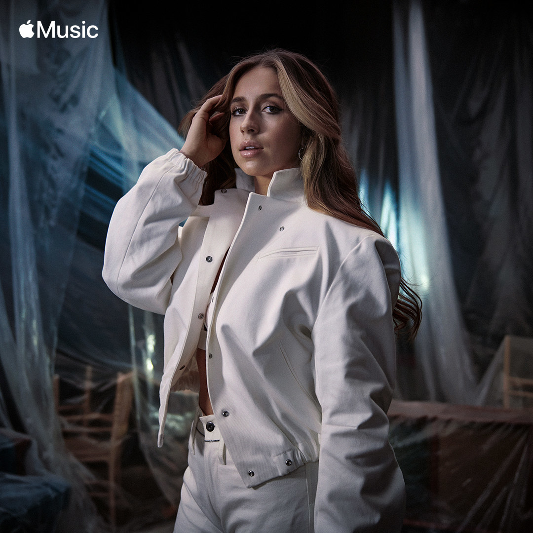 Tate posing in a room with overturned chairs covered in sheer fabric as she wears a matching jacket and pants combo for a promotional photo