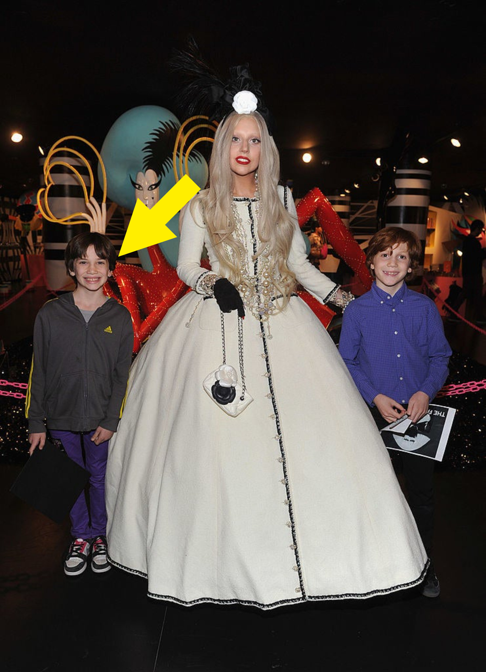 Lady Gaga is wearing a ballgown and matching floral headpiece as she poses for a photo with two young children, one of whom has an arrow pointing at him identifying him as Bill Gates