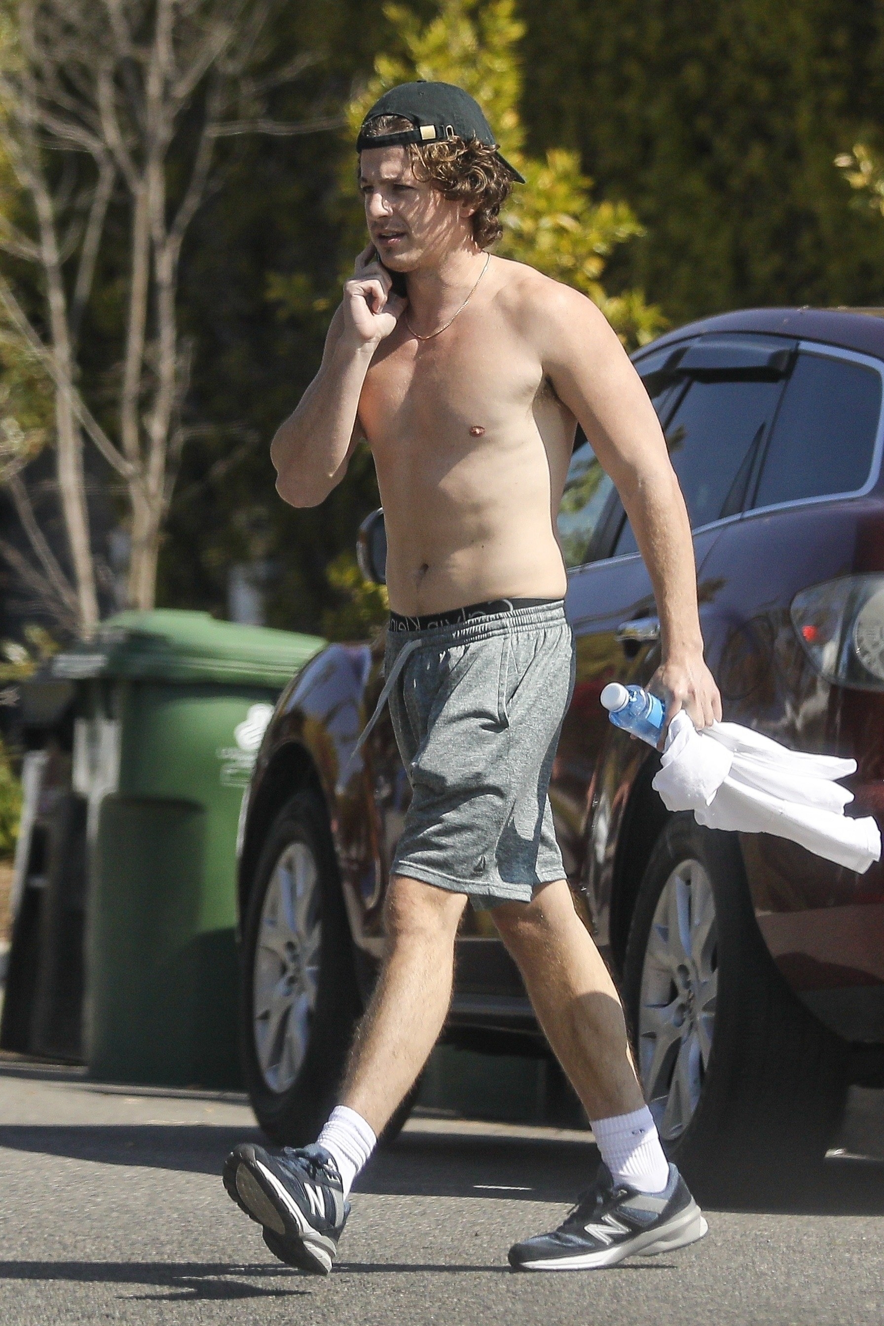 Another photo of shirtless Charlie wearing shorts and sneakers and holding a phone, T-shirt, and cellphone