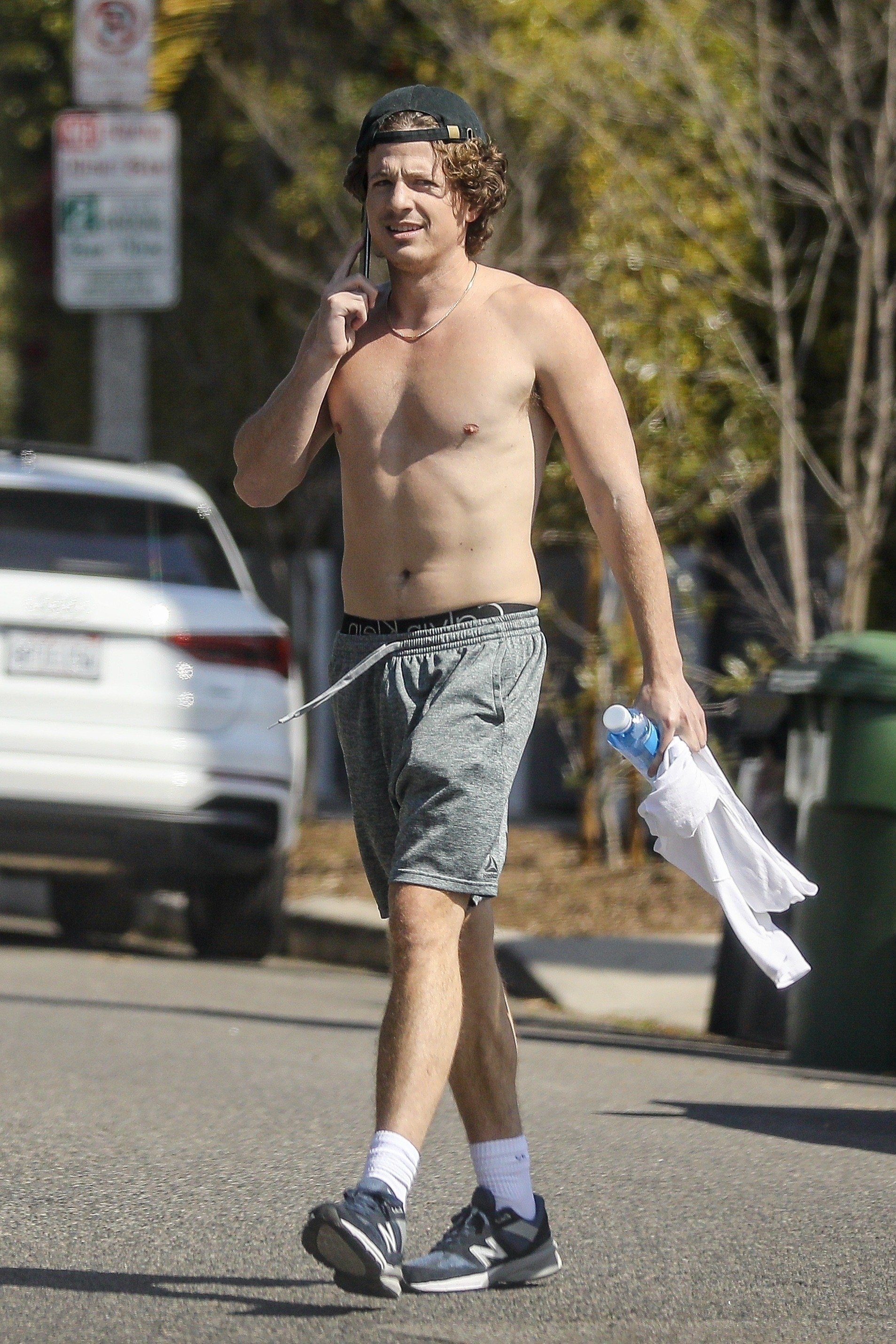 Shirtless Charlie walking, wearing shorts and sneakers, and holding a phone, T-shirt, and cellphone