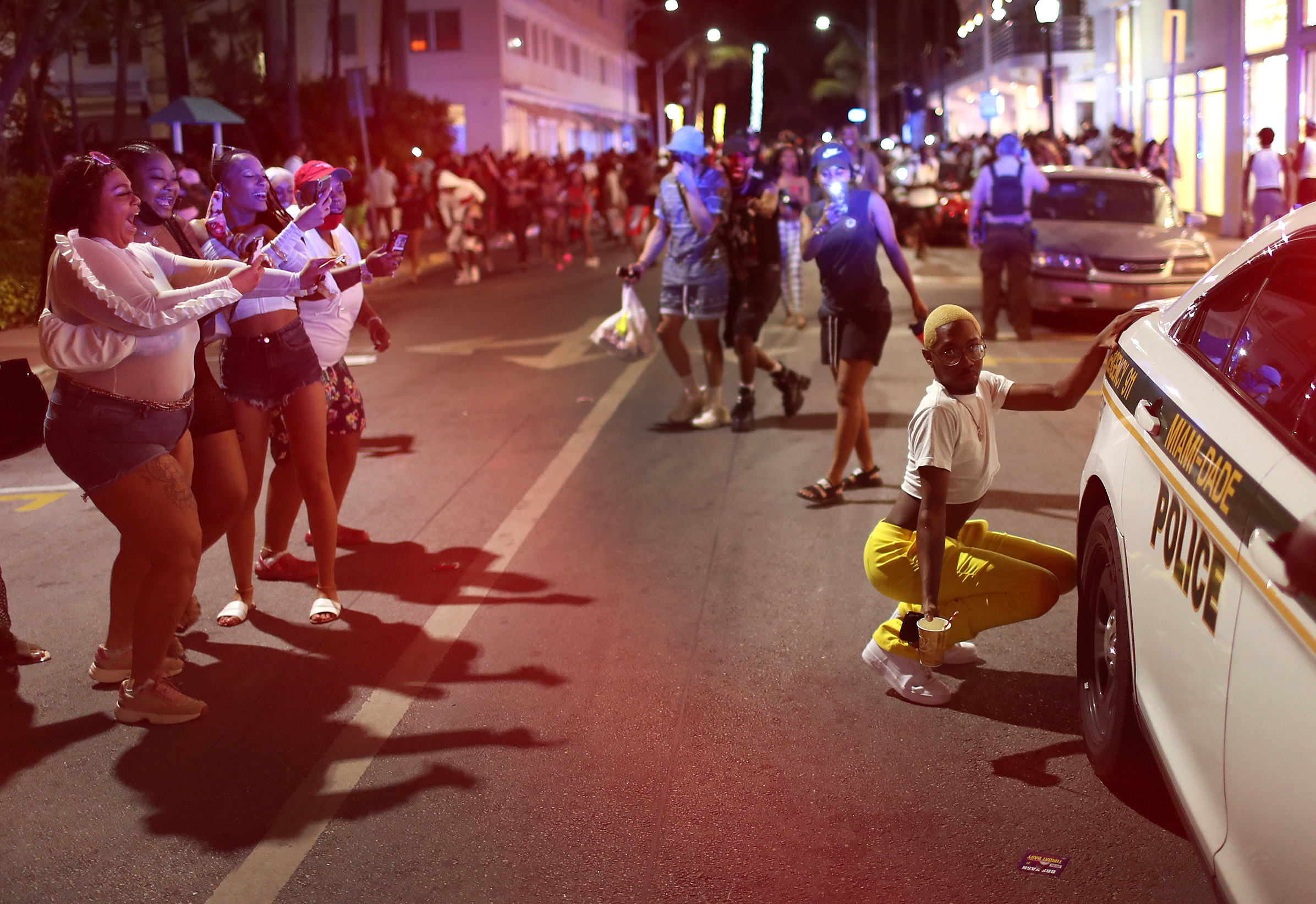 A person squats next to a cop car while women take a photo of them on a crowded street