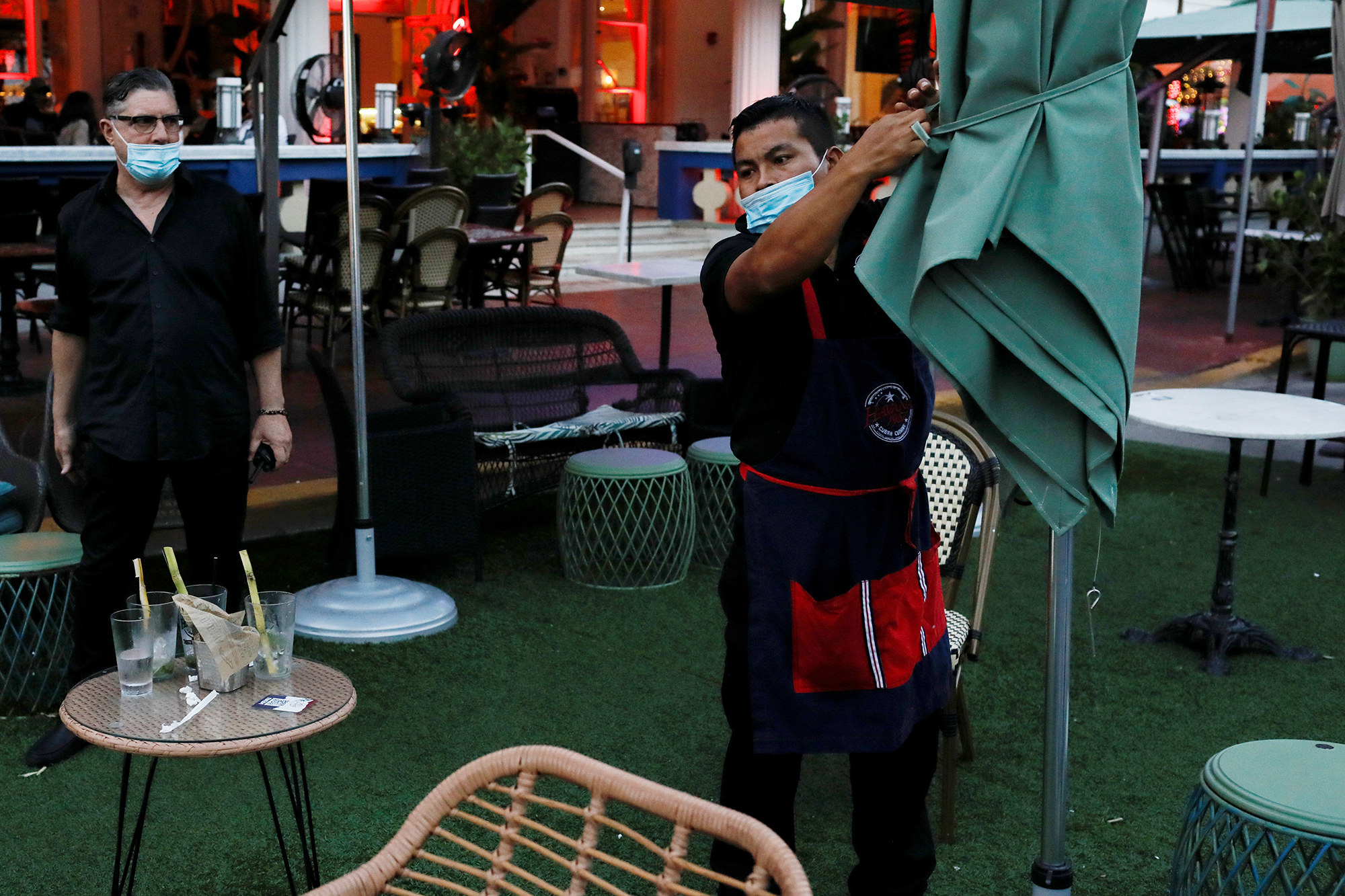 A man wearing a mask packs up an umbrella at an outdoor dining setup 