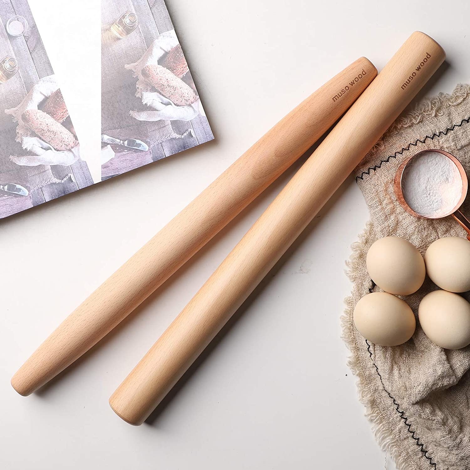 two muso wood tapered french rolling pins on a marble counter next to a few eggs and a measuring cup full of flour