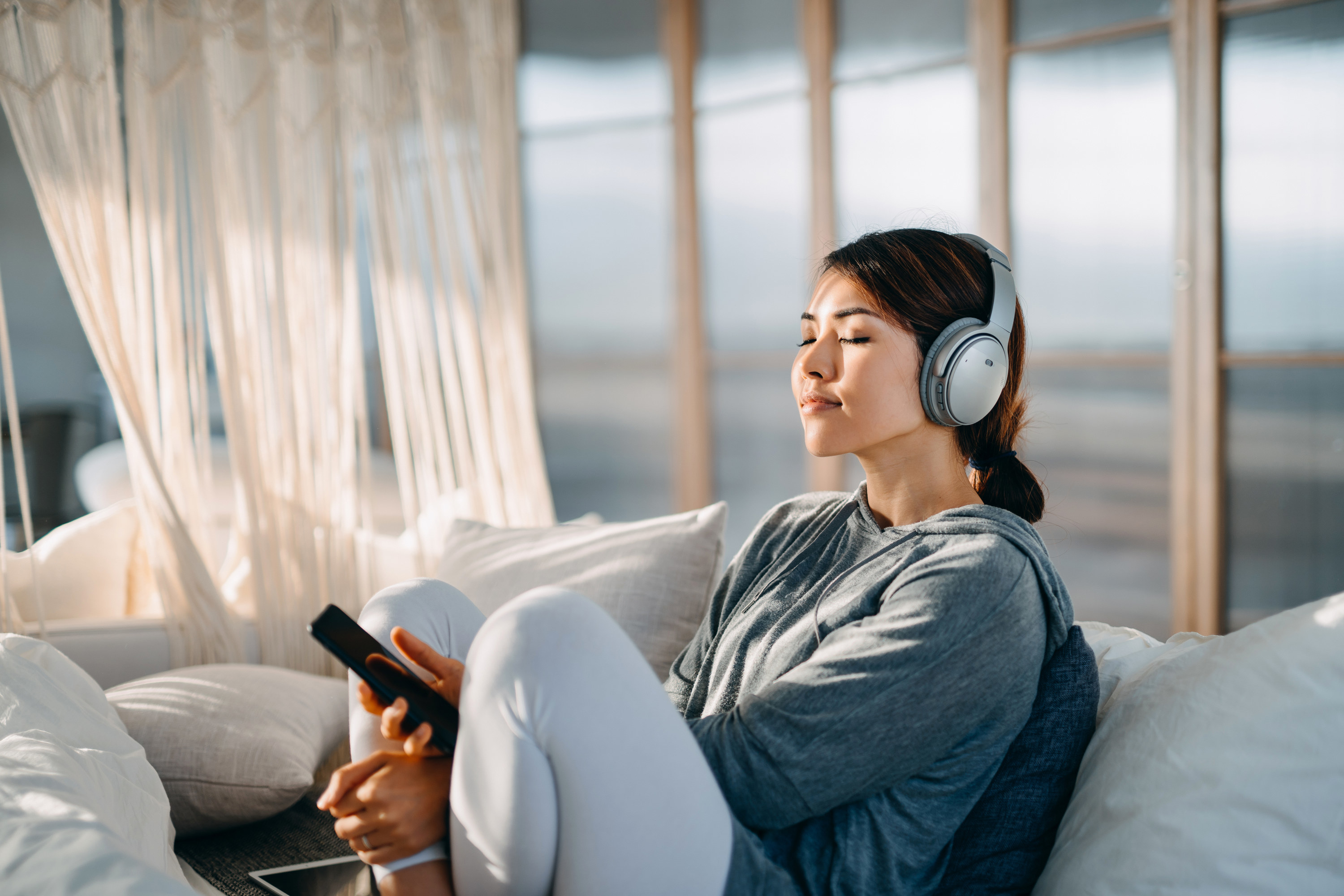 Younger woman listening to music on headphones, while sitting on a white sofa in breezy-looking room