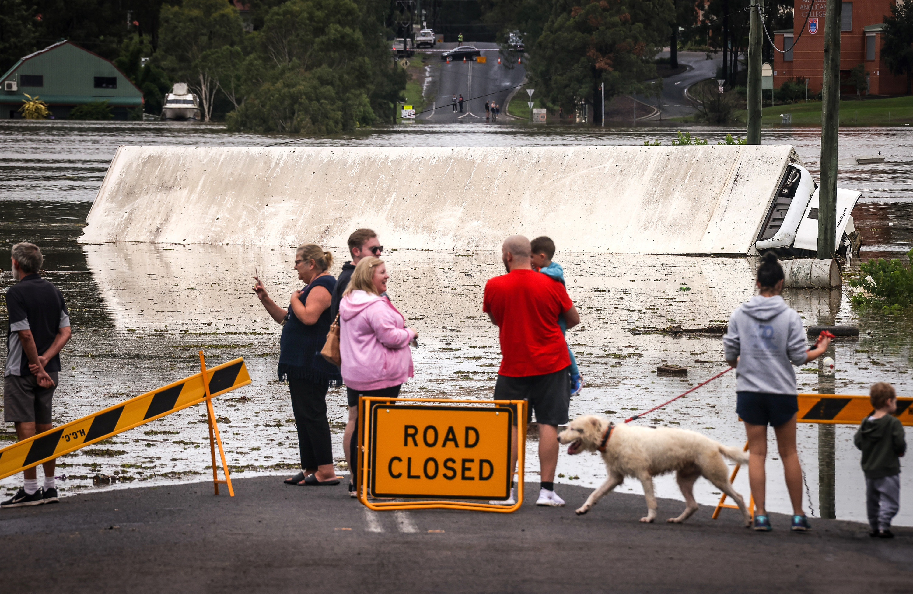 People gather at the end of a flooded area, with road closed signs, to take pictures of a tipped over truck