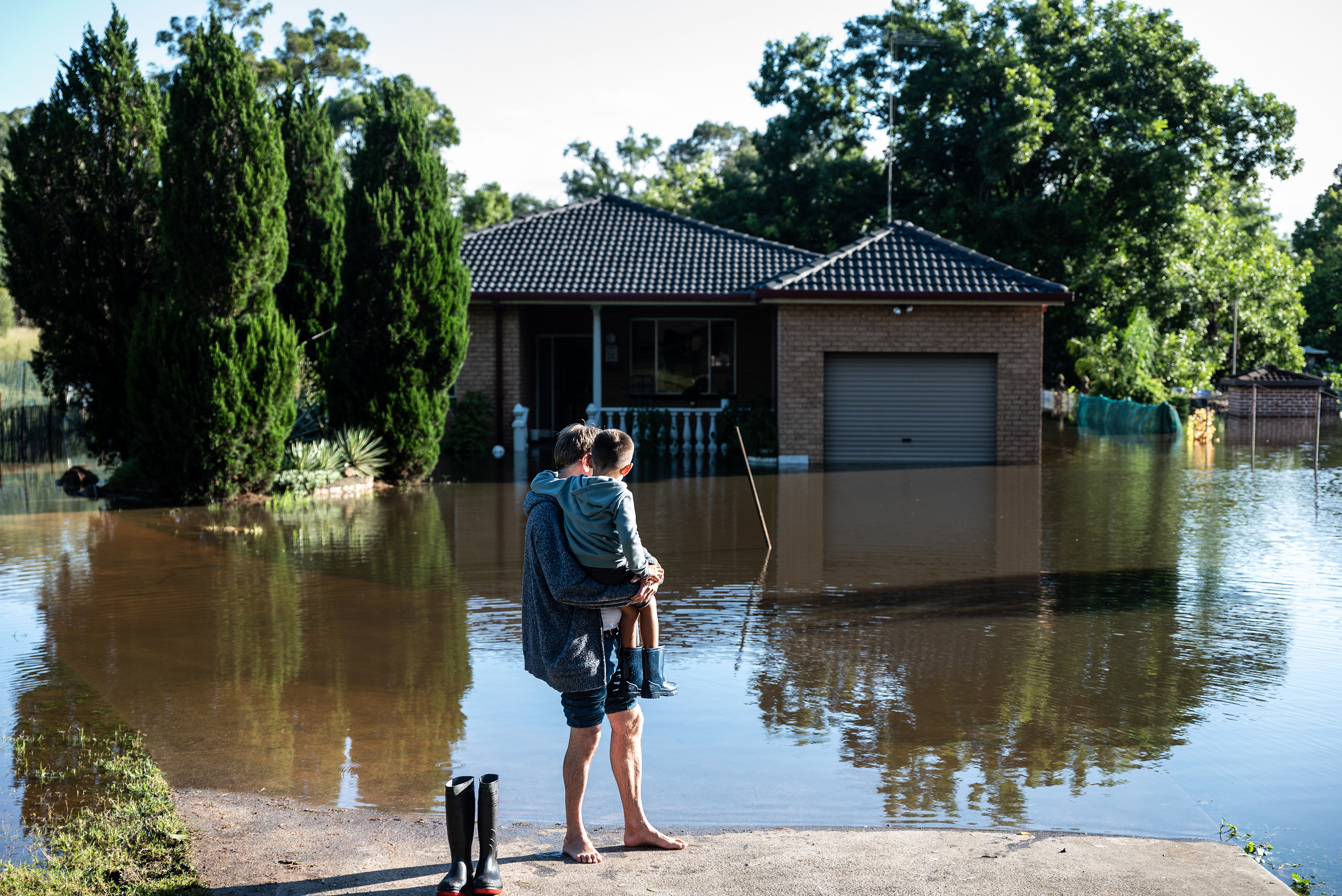 A father and son stand on dry land in front of a flooded home