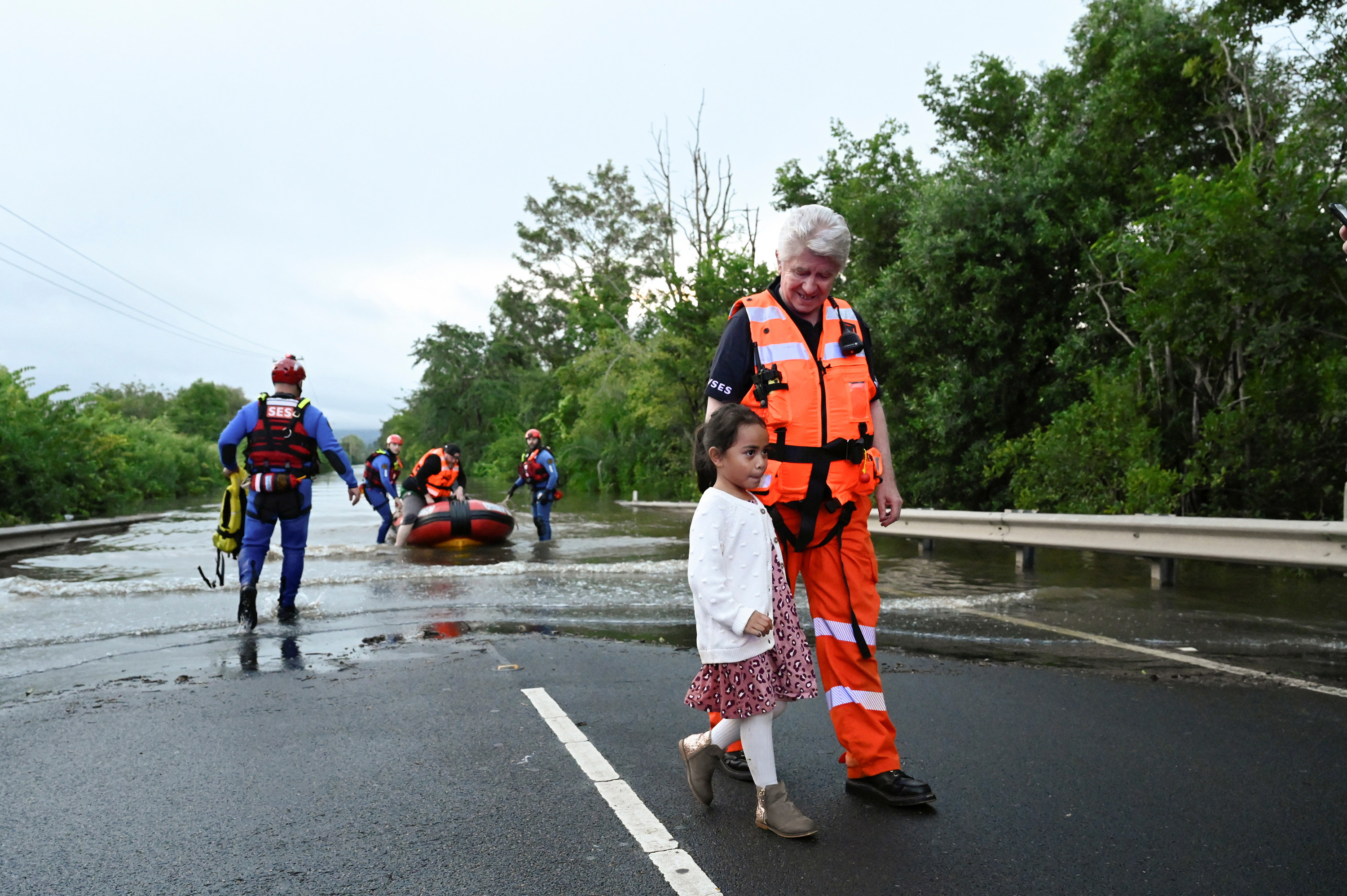 A woman in a drysuit escorts a young girl away from floodwater, behind them two workers assist a man getting out of the boat