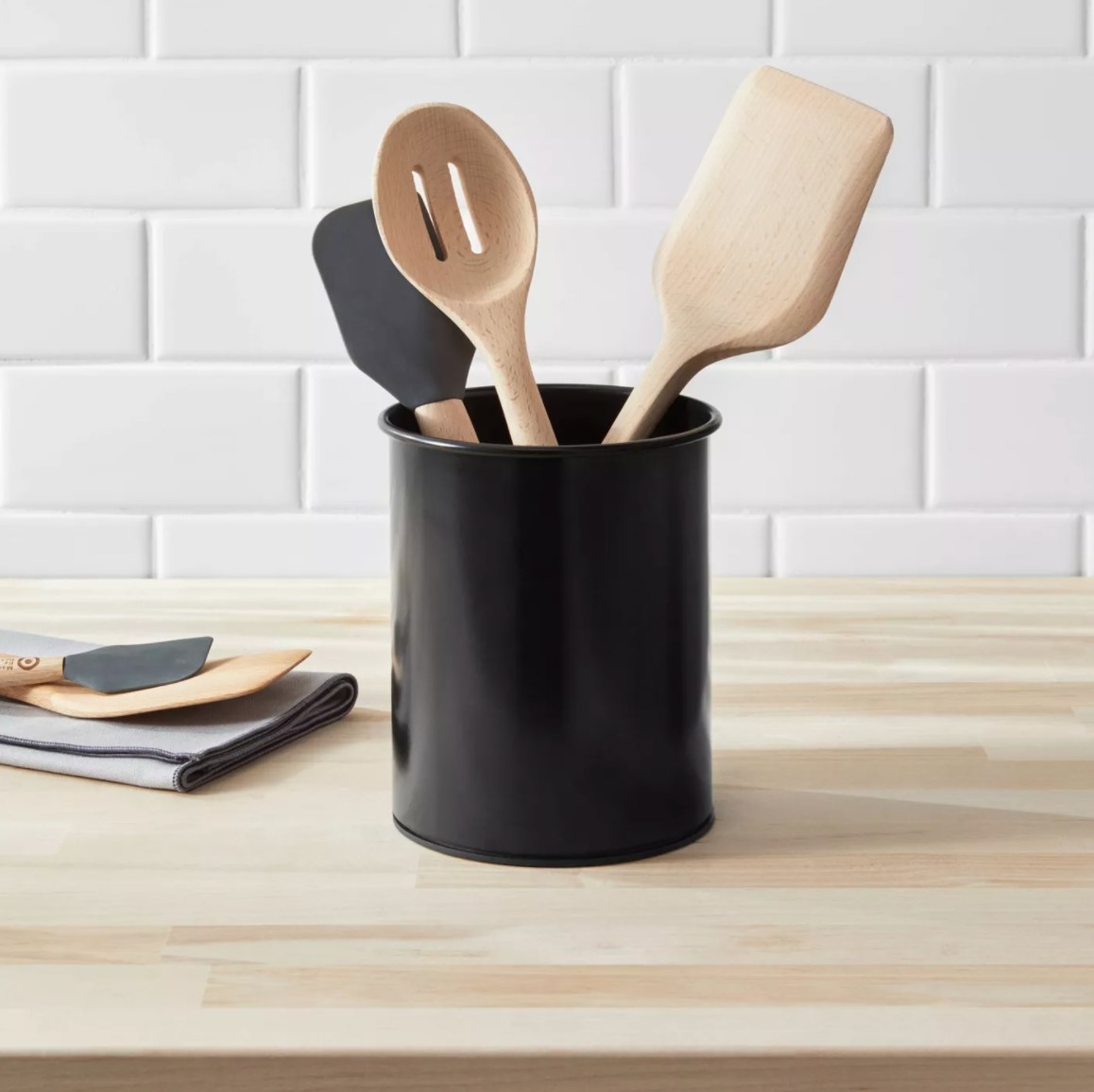 The stainless steel storage container in black holding a rubber spatula, slotted spoon, and wooden spatula