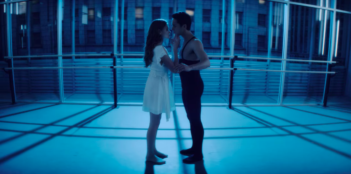 A woman in a white dress and a man in a black leotard and black tights embrace each other in a dance studio.