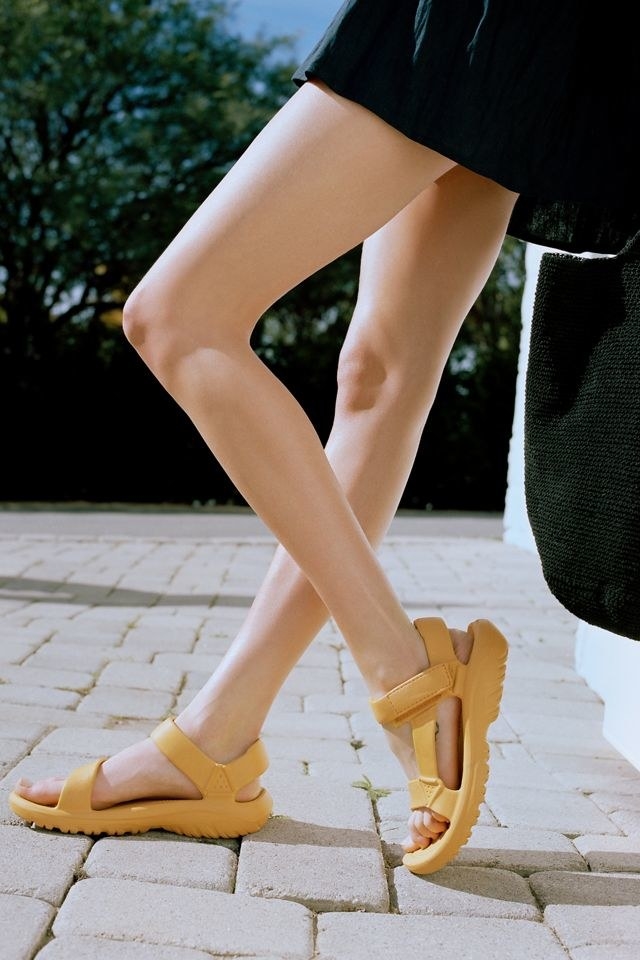 model wears yellow Teva sandals with a cushy footbed on stone pavement