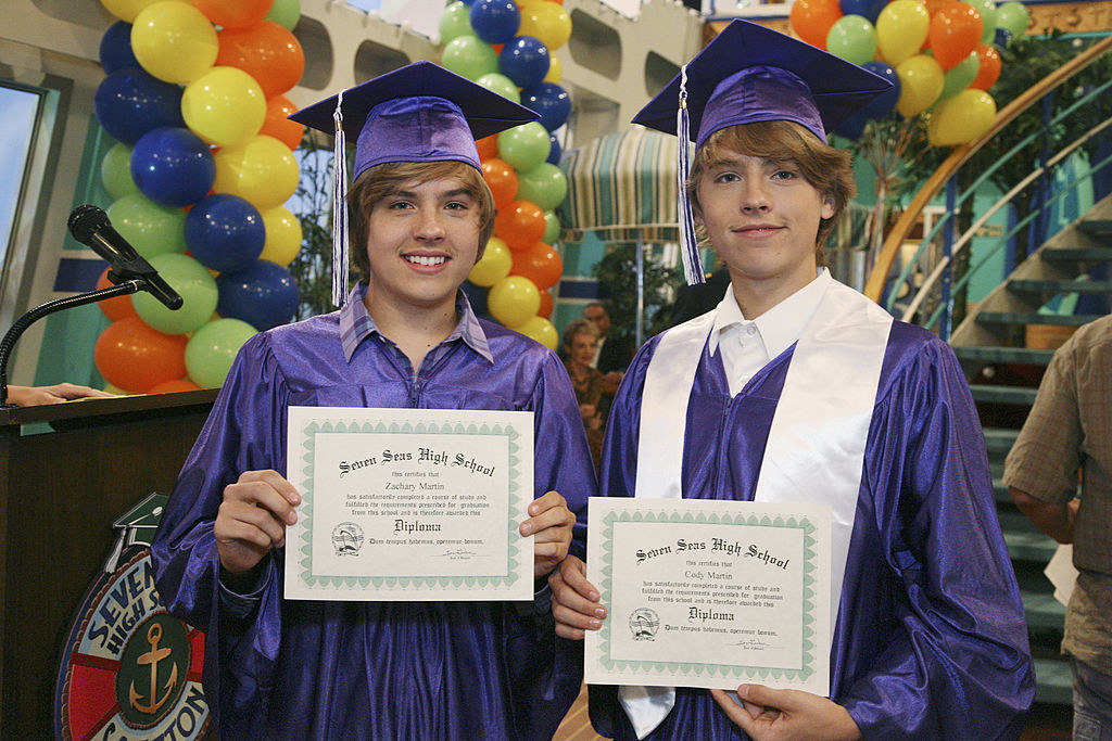 Cole Sprouse (R) and Dylan Sprouse wearing graduation gowns and holding up high school diplomas in a scene for the Suite Life