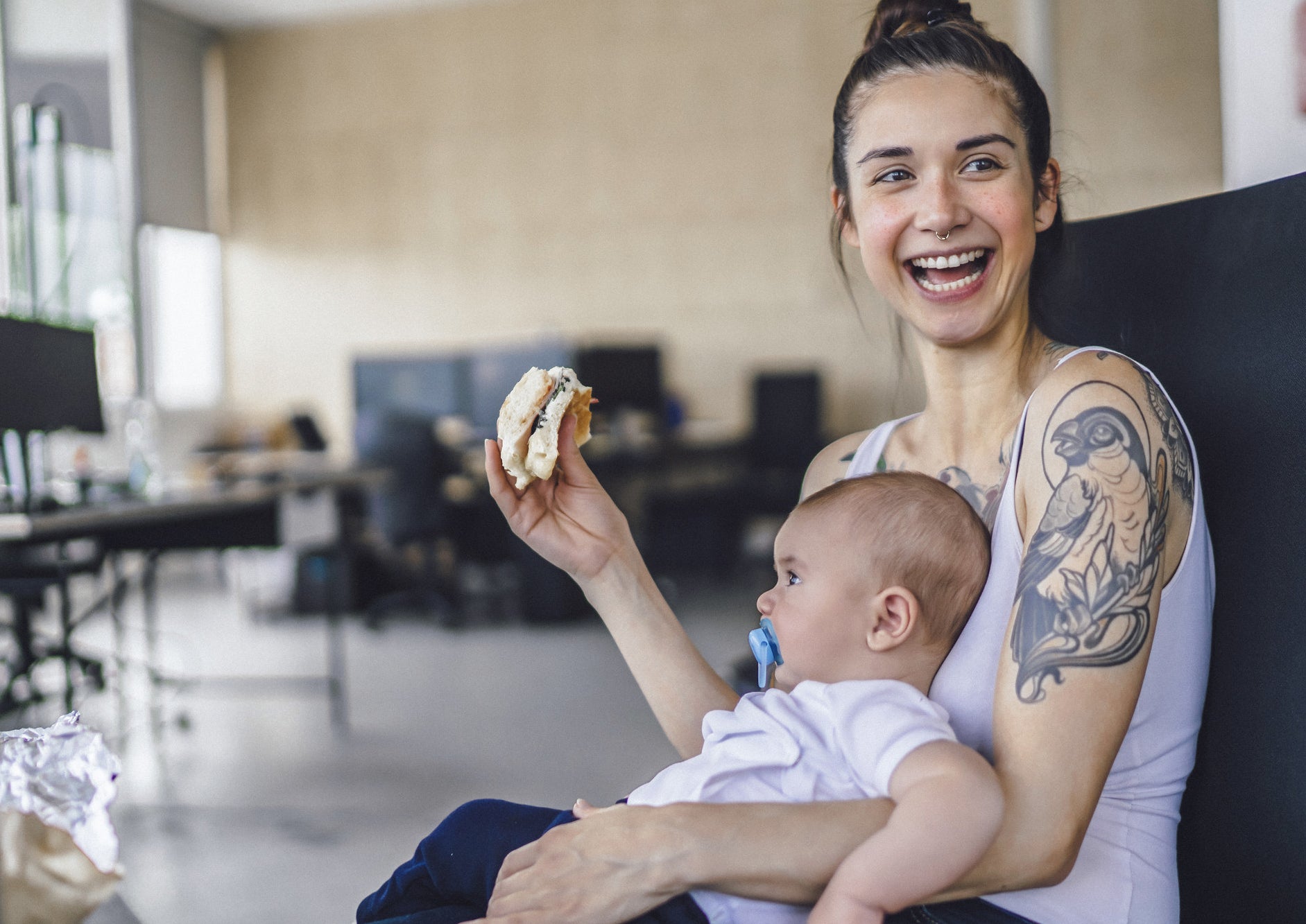 A smiling mom with tattooes holds her baby and a sandwich