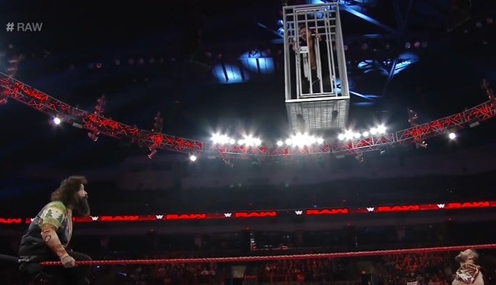 Wrestler dangling above the ring in a shark cage.