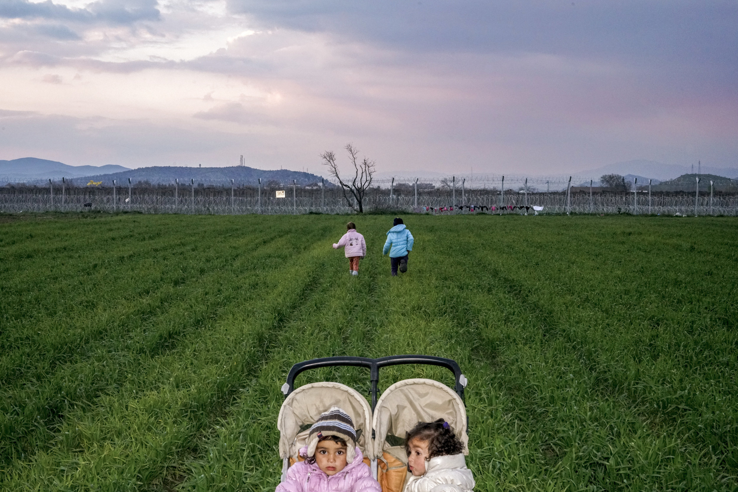 two small children in a push chair while their mothers walk in a field behind barbed wire