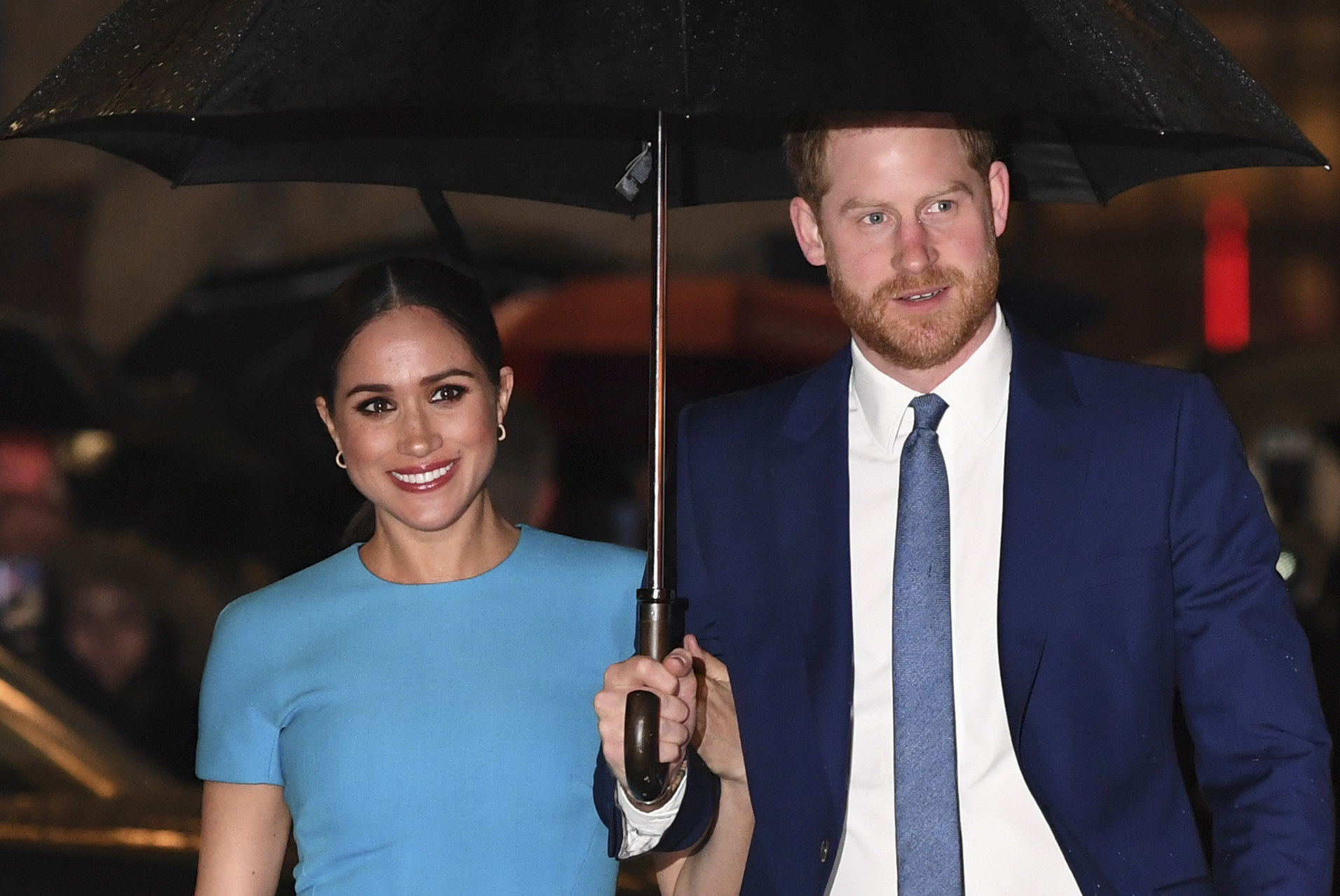 Meghan smiles next to Prince Harry while standing under an umbrella