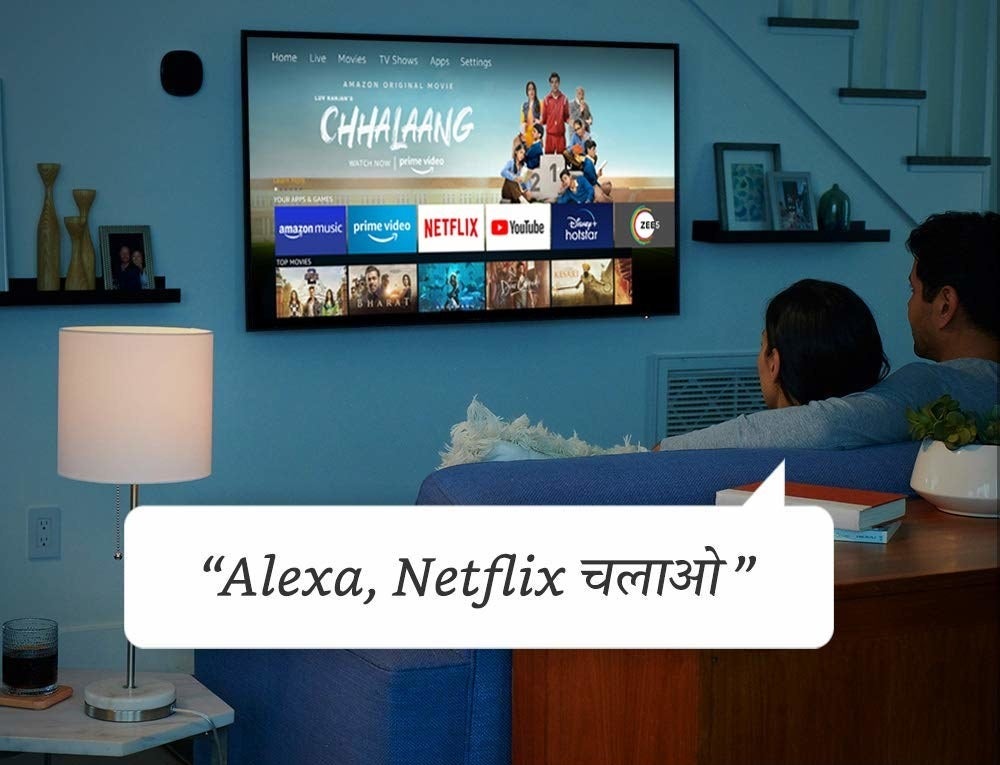 Two people in front of their TV, with a dialog box saying, “Alexa, Neflix chalao/open] Netflix.”