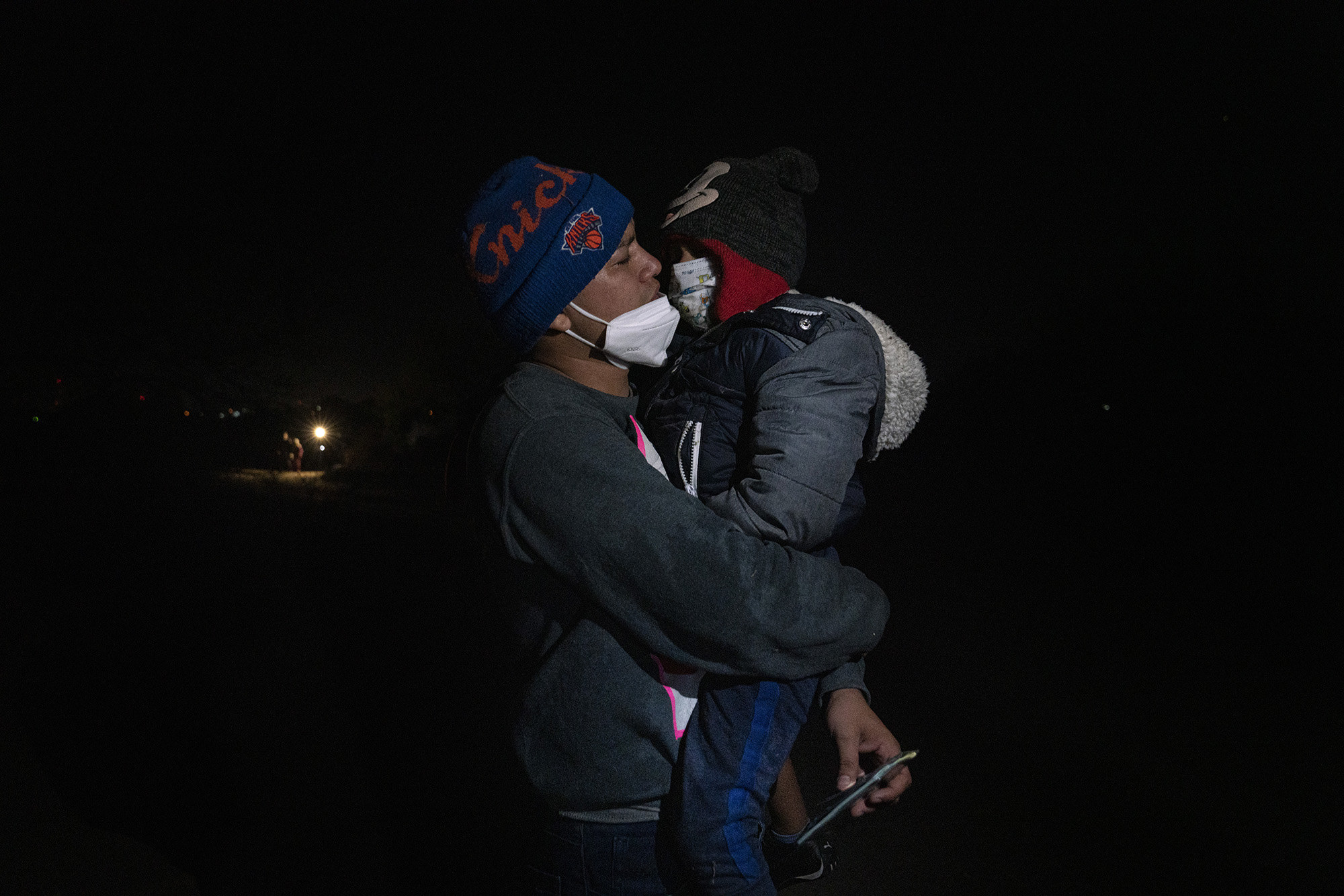 A father holds his son, both dressed in beanies, face masks, and warm clothing at night