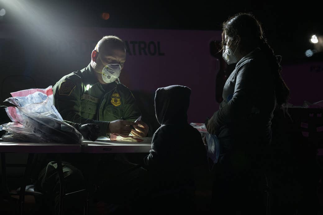 A mother and child stand by a table, where a sitting Border Patrol agent holds a pen and document in his hands