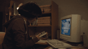 Pen15 GIF of Maya dialing a &#x27;90s portable phone in front of a desk top computer