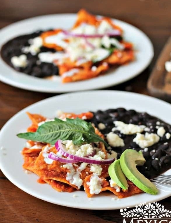 Chilaquiles rojo topped with cheese, avocado, onion, and plated with black beans. 