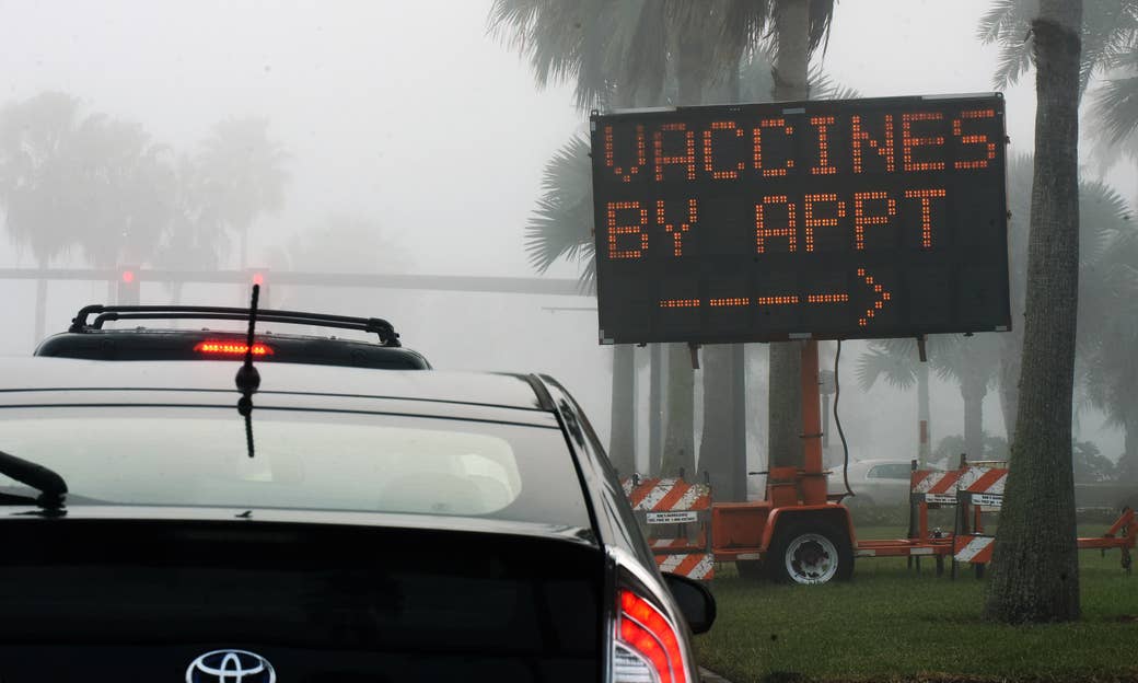 A large roadside sign reads &quot;Vaccines by appointment&quot; with an arrow pointing to the left as cars line up
