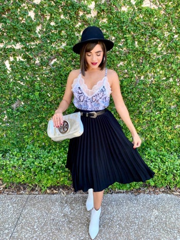 reviewer wearing the black pleated skirt with a cami, white boots, and fedora