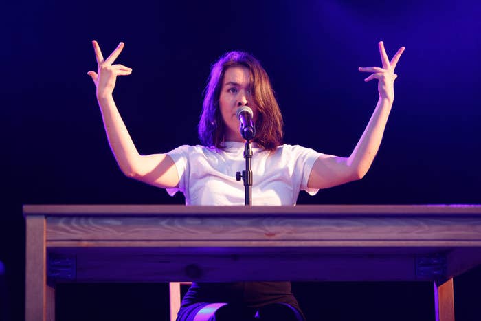 a woman wearing a t-shirt stands above a keyboard with her hands in the air, fingers spread. she is singing into a microphone