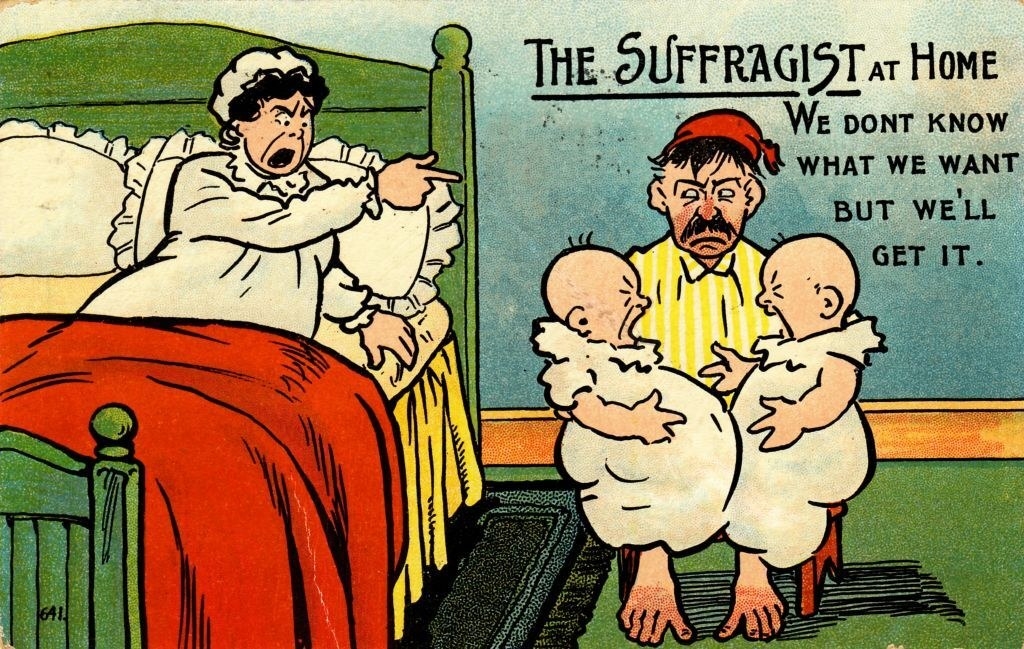 A woman yelling at her husband, who&#x27;s holding two babies, with the caption, &quot;The Suffragist at home, we don&#x27;t know what we want but we&#x27;ll get it&quot;