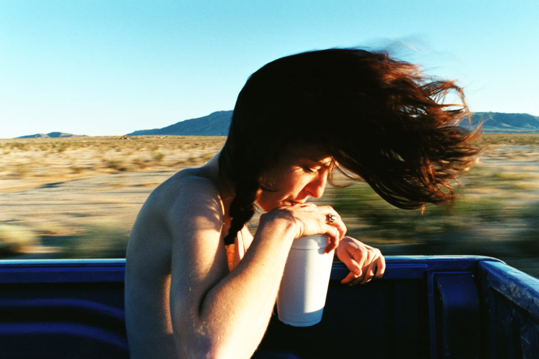 A naked young person sips out of a styrofoam cup in the back of a moving truck while their hair flies and the landscape whizzes by