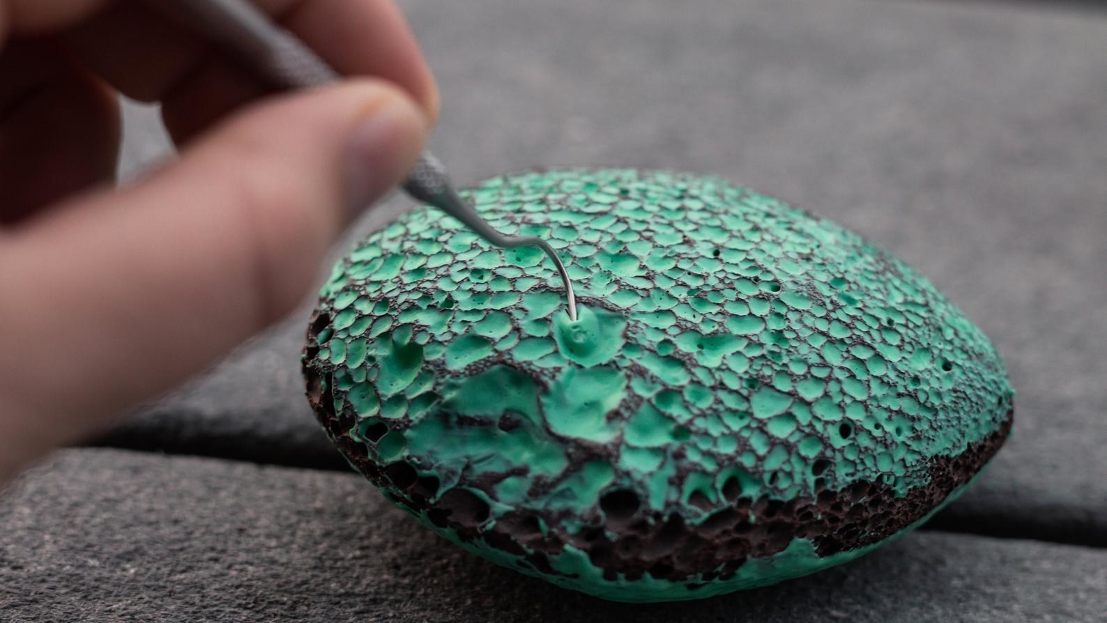 A model using a tiny hook to pick blue paint out of a lava rock with tiny holes