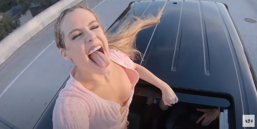Stefani standing through the sunroof of a moving car and sticking her tongue out with glee